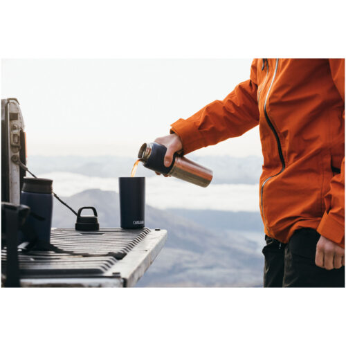 Eine Person in einer orangefarbenen Jacke gießt ein Getränk aus einer Thermoskanne in einen Becher auf einer Heckklappe mit Blick auf die Berge.