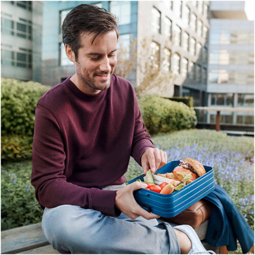 Mann sitzt lächelnd im Freien und hält eine blaue Lunchbox mit einem Sandwich und Gemüse.