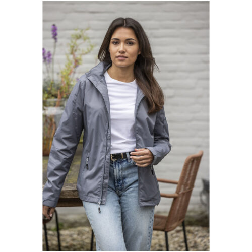 Frau in grauer Jacke und Jeans steht im Freien an einem Holztisch vor einer hellen Backsteinwand.