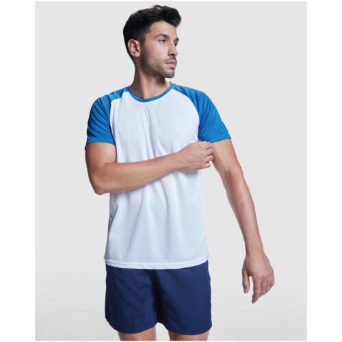 Ein Mann in einem weiß-blauen Sportshirt und Shorts steht vor einem schlichten hellen Hintergrund.